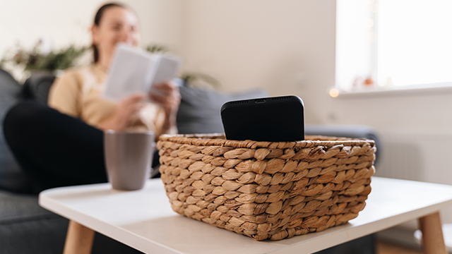 Een gevlochten mand met een zwarte telefoon staat op een witte tafel, met een vage vrouw die een boek leest op de achtergrond.