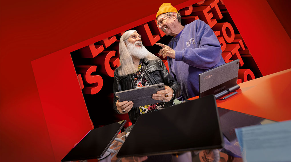 Twee mannen staan achter bureaus met laptops tegen een rode achtergrond met de tekst "LET'S GO".