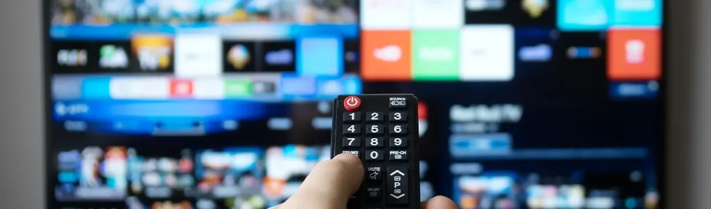 A hand holds a remote control in front of a TV with a colorful interface.