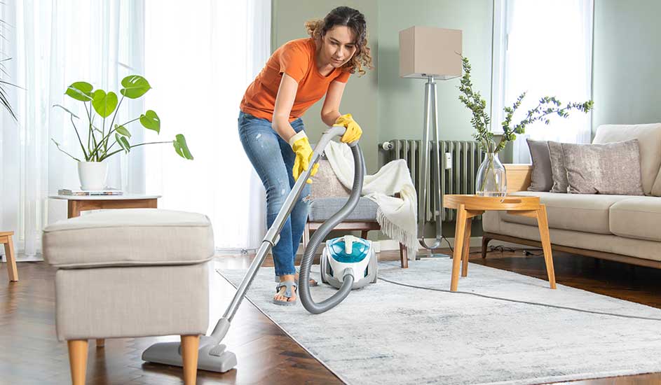 Een vrouw stofzuigt een lichte woonkamer, draagt gele handschoenen, terwijl planten en houten meubels de ruimte gezellig en opgeruimd maken.