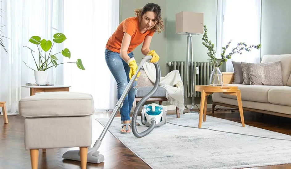 A woman hoovers a bright living room, wearing yellow gloves, as plants and wooden furniture keep the space airy, tidy and homely.