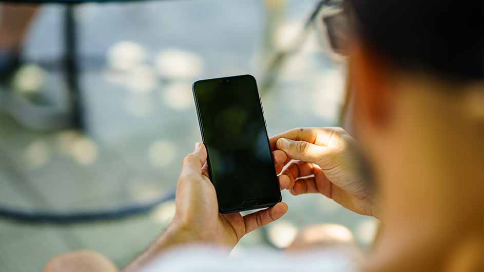 Een persoon houdt een zwarte smartphone vast. De achtergrond is vaag met elementen van een tafel en stoelen.