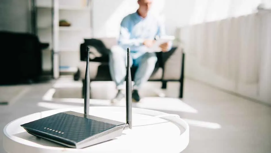 Een zwarte router met twee antennes staat op een ronde, witte tafel. Op de achtergrond zit een persoon op een grijze bank.