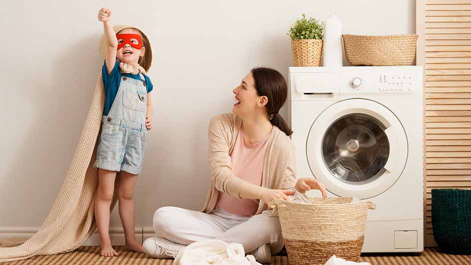 Een kind verkleed als superheld staat naast een lachende vrouw met een wasmand in een kamer met een wasmachine.