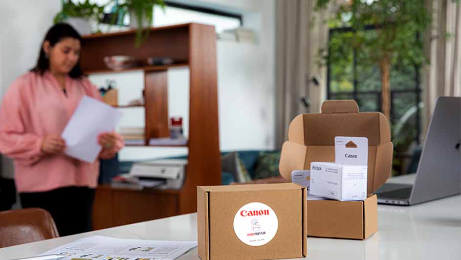 Bruine Canon inktcartridge dozen op een witte tafel, open doos toont cartridges. Vrouw met papier en laptop in de achtergrond.