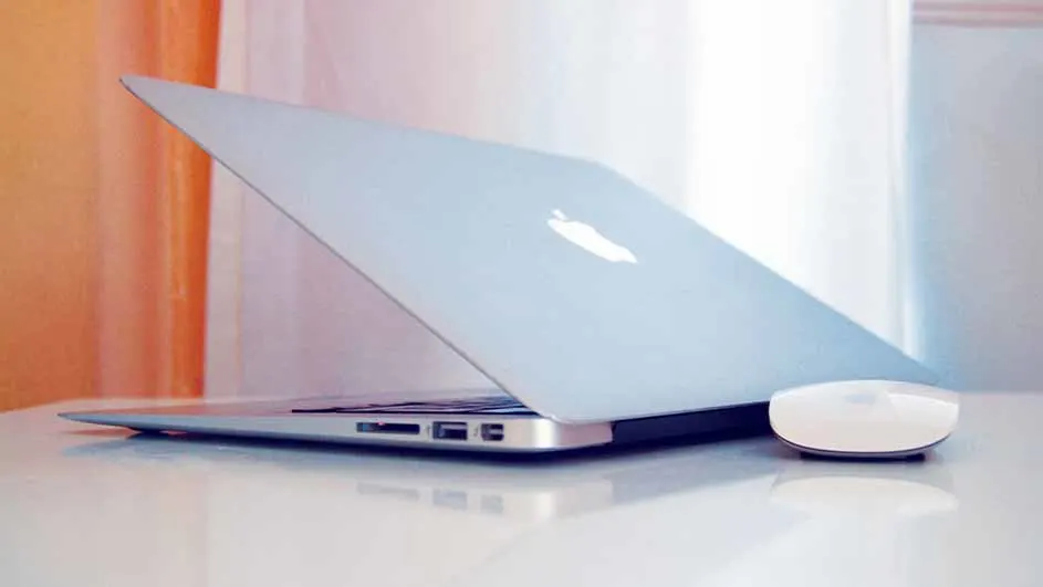 Een zilverkleurige laptop staat open op een witte tafel, met een witte draadloze muis ernaast. Achtergrond: gordijnen in wit en oranje.