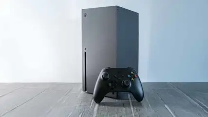 Een grijze Xbox Series X staat op een houten vloer, met een zwarte controller ervoor. De achtergrond is lichtblauw.