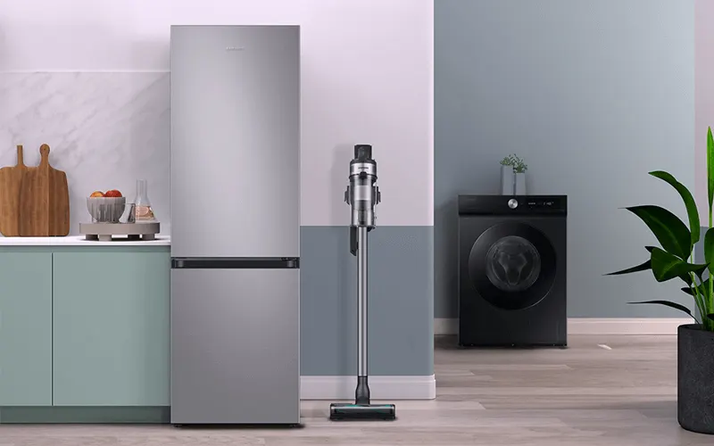 Keuken met een zilverkleurige koelkast, een staande stofzuiger, een zwarte wasmachine en groene planten.