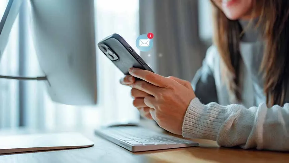 A person holds a phone with a new email notification. A computer and keyboard are on a desk.