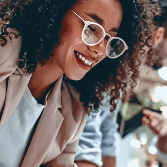 Close up of a smiling woman with glasses leaning over a desk, looking focused and enthusiastic while collaborating with colleagues.