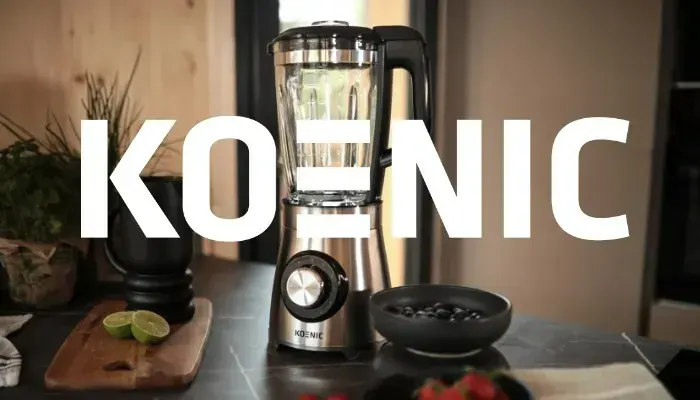 A stainless steel Koenig blender sits on a counter with a black bowl of blueberries, limes on a wooden board, and plants.