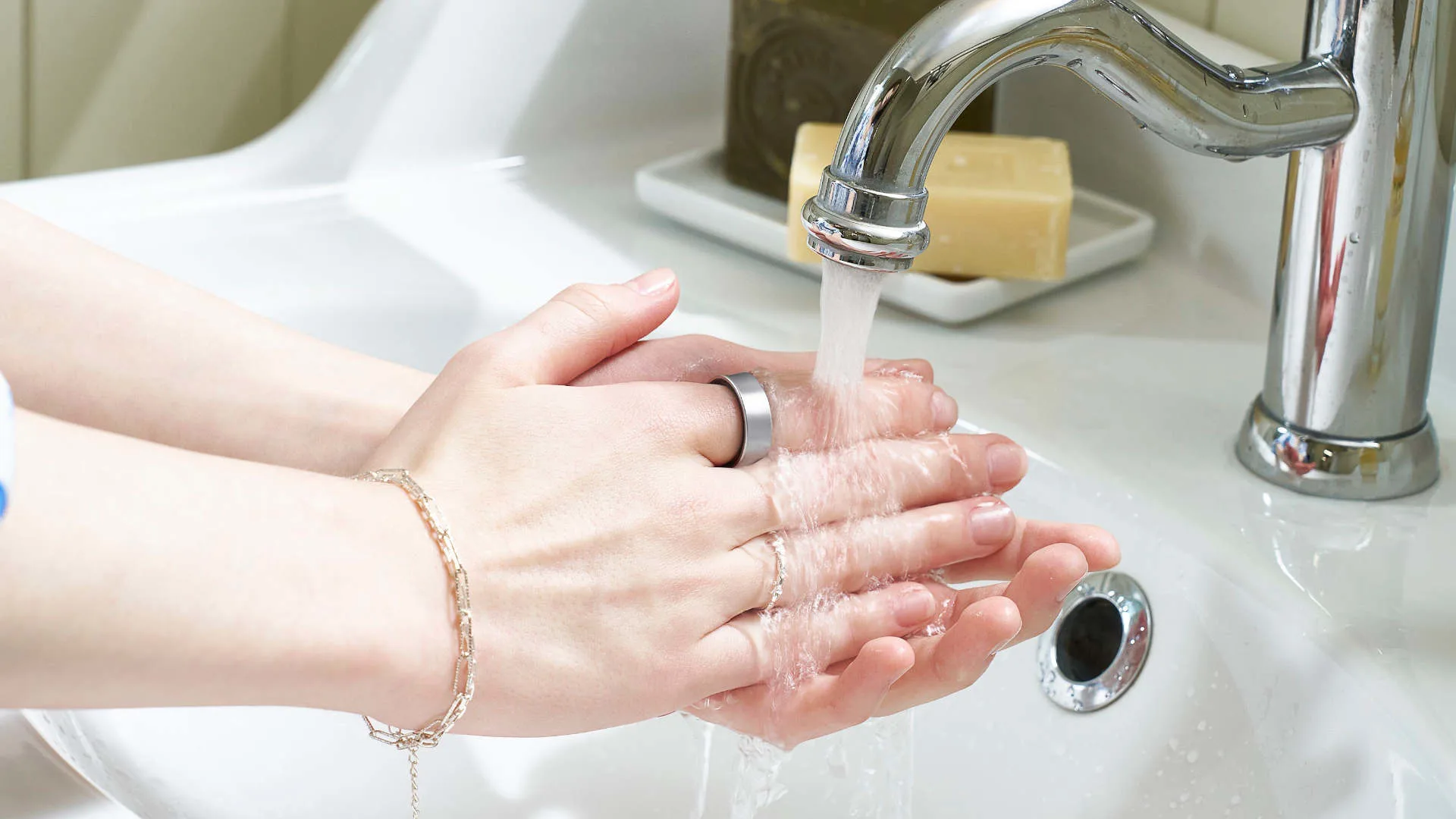 Twee handen met zilveren ringen wassen zich onder een chromen kraan in een witte wasbak. Op de achtergrond staat een zeepje.