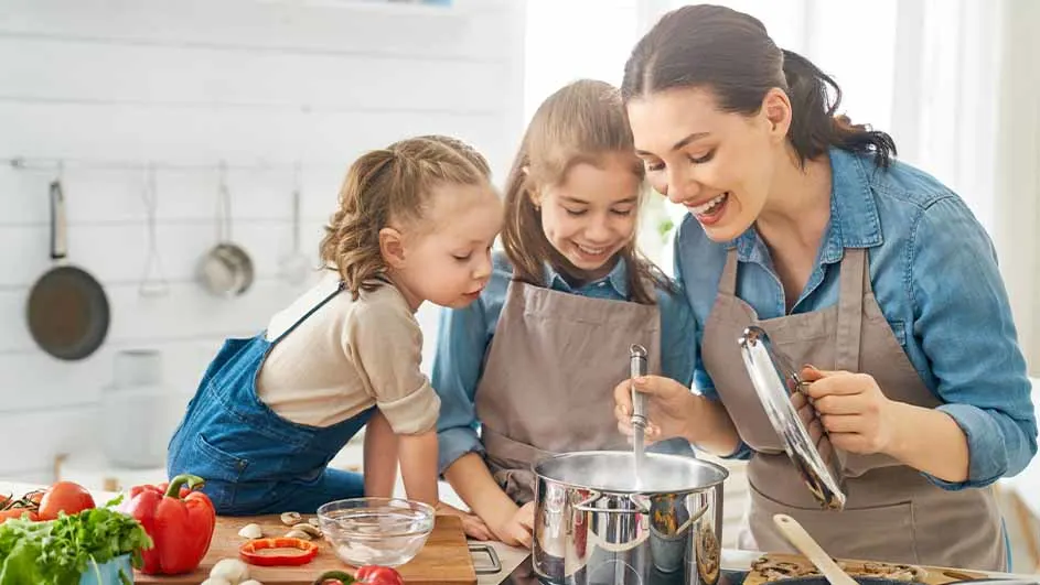 Een vrouw en twee meisjes in schorten koken samen. Een pan staat op het fornuis, omringd door groenten.
