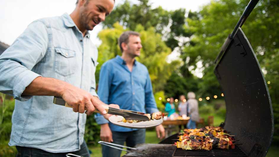 Twee mannen bij een barbecue met spiesen. Een man gebruikt een tang. Achtergrond: groene bomen en mensen bij een tafel.