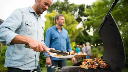 Twee mannen bij een barbecue met spiesen. Een man gebruikt een tang. Achtergrond: groene bomen en mensen bij een tafel.