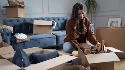 Vrouw pakt mok uit doos in kamer vol verhuisdozen. Op de achtergrond een blauwe bank en een kamerplant.