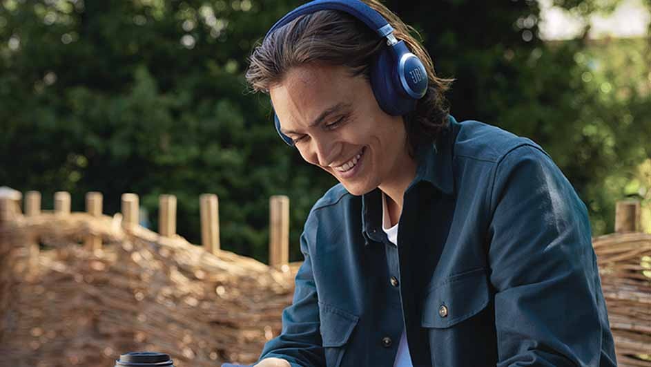 A person smiles, wearing blue headphones and a button-down shirt. A cup sits on a wooden table with a blurred green background.