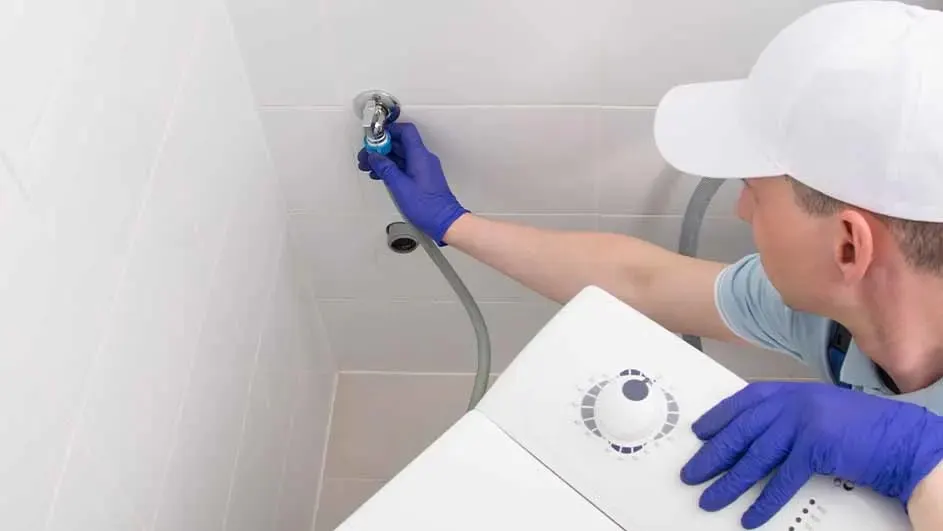 Een loodgieter met witte pet en blauwe handschoenen sluit een witte wasmachine aan op een waterkraan in een betegelde ruimte.