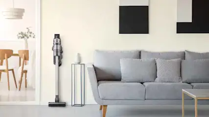 Een stofzuiger staat aan de muur naast een grijze bank met kussens en een bijzettafel in een woonkamer.