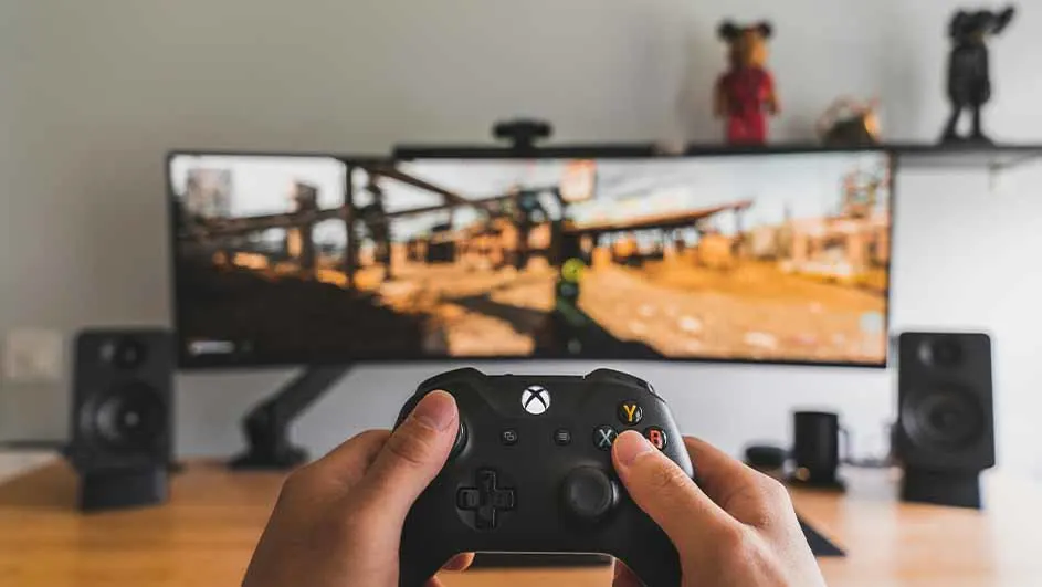Twee handen houden een zwarte Xbox-controller vast voor een groot scherm met een spel. Speakers staan aan weerszijden, een plank erboven.