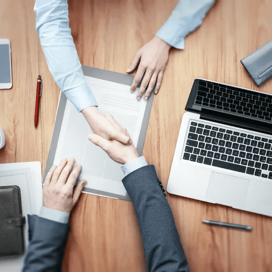 Top view of a wooden meeting table with laptops, papers and pens, as two business people exchange a firm, professional handshake.