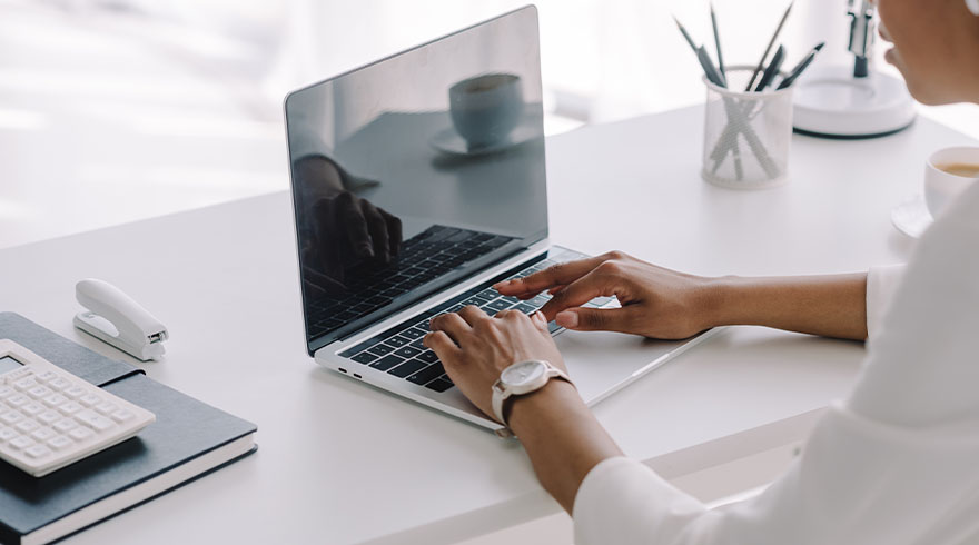 Een persoon typt op een laptop op een wit bureau met een rekenmachine, nietmachine en kopje.