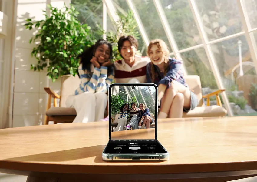 Drie mensen zitten op stoelen voor een glazen wand, gefotografeerd door een opengeklapte smartphone op een houten tafel.