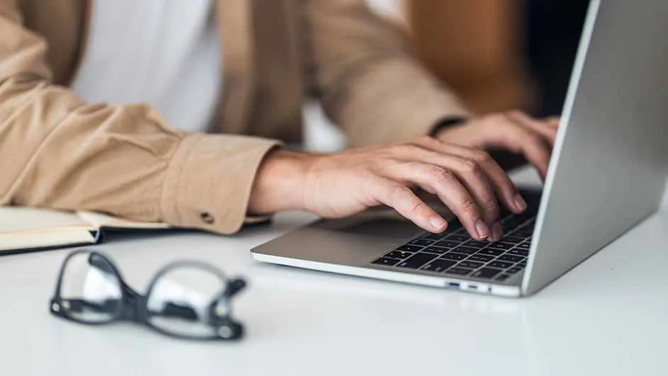 Man typt op laptop, met notitieboek en bril op een wit bureau.