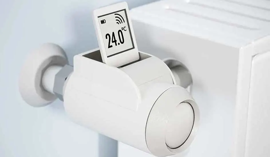 White smart thermostat on a radiator displays 24.0 degrees Celsius. The device shows battery and signal icons.