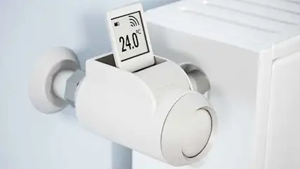Een witte thermostaat is bevestigd aan een radiator met een display dat 24 graden toont.
