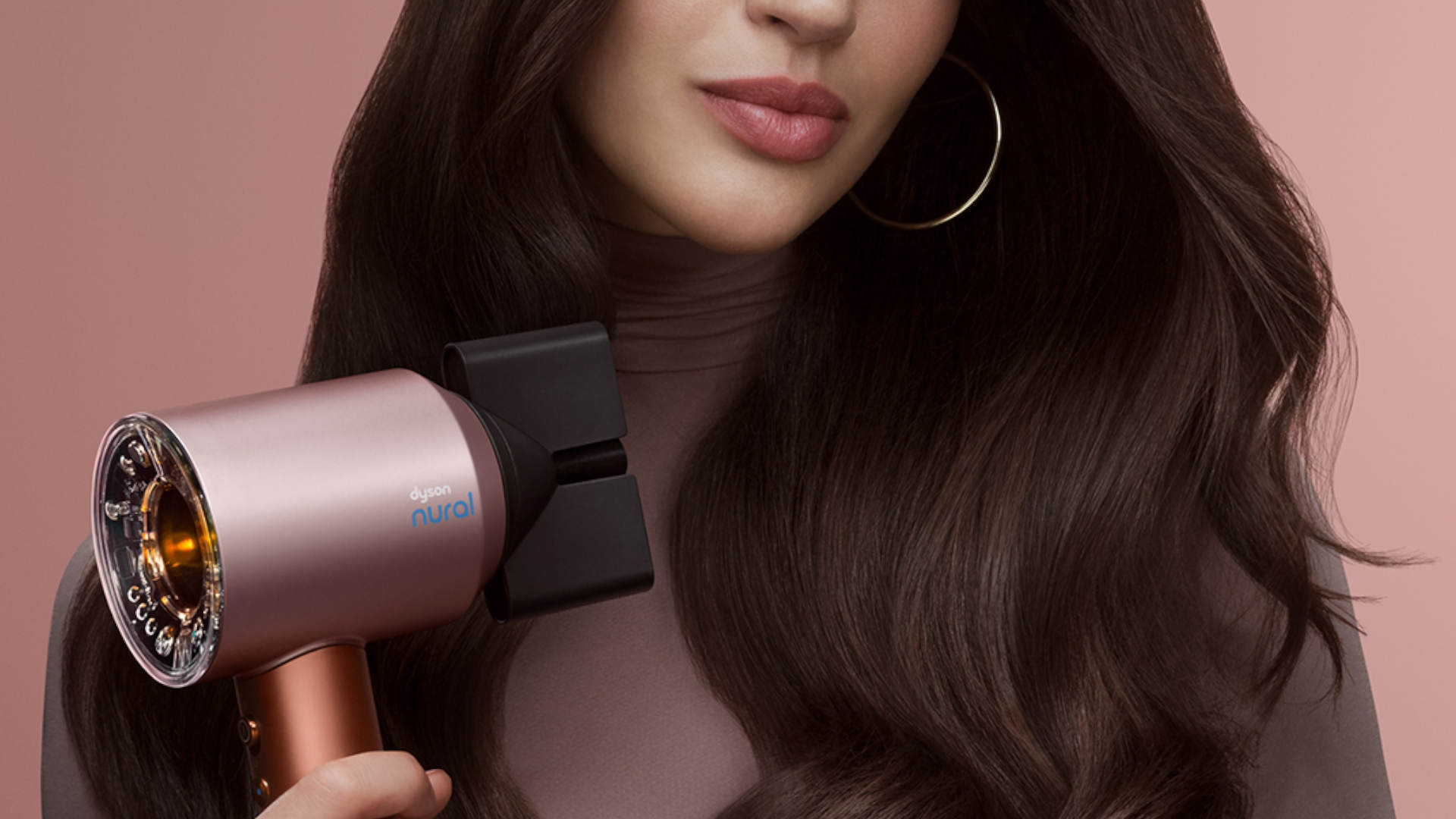 Vrouw met lang, glanzend bruin haar houdt een roségouden föhn vast tegen een roze achtergrond.