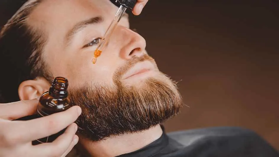 Een man krijgt baardolie toegediend met een pipet, terwijl een bruin flesje wordt vastgehouden. De achtergrond is donker.