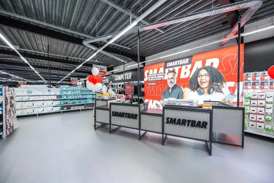 Interieur van een MediaMarkt winkel met schappen vol producten, een Smartbar balie en reclameposters.