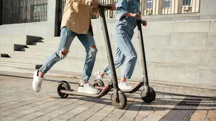 Two people riding scooters on a brick path, dressed in casual clothing and white sneakers, with a building visible in the background.