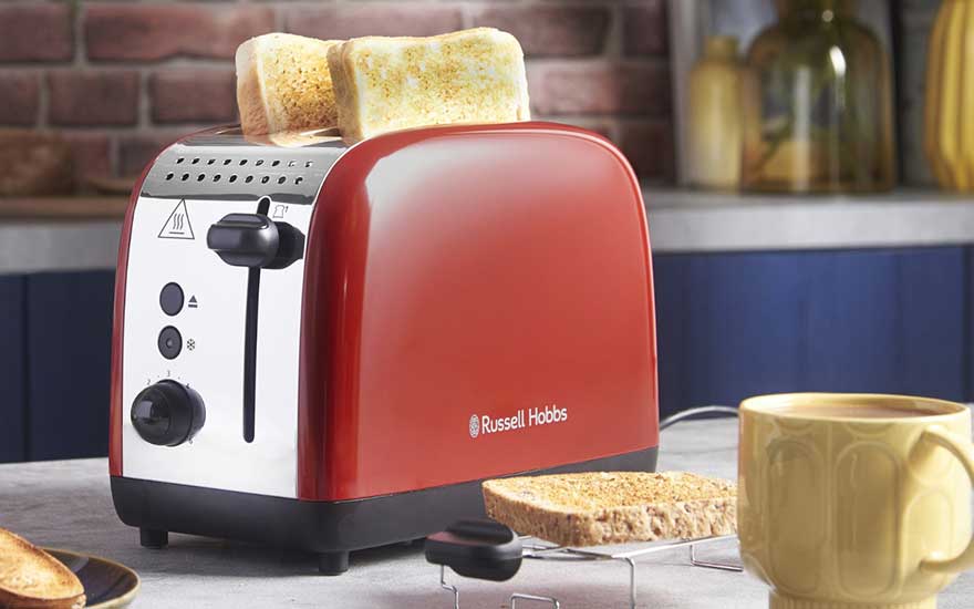 Een rode Russell Hobbs broodrooster met twee goudbruine sneetjes brood, omringd door geroosterd brood op tafel en een gele mok met warme drank.