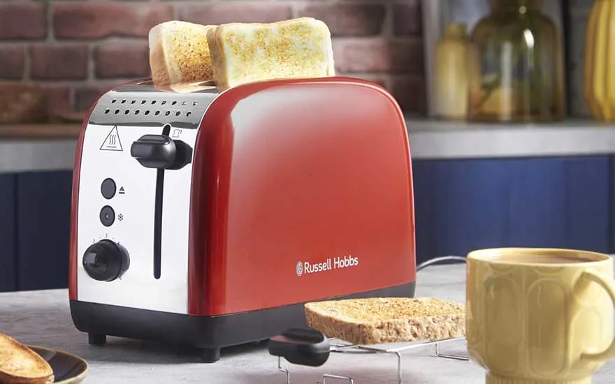 Een rode Russell Hobbs broodrooster met twee goudbruine sneetjes brood, omringd door geroosterd brood op tafel en een gele mok met warme drank.