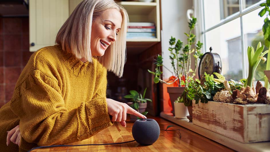 Een vrouw in een gele trui bedient een slimme speaker op een houten tafel bij een raam met planten.