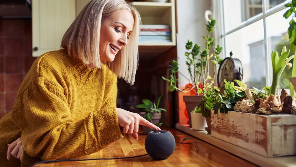 Een vrouw in een gele trui bedient een slimme speaker op een houten tafel bij een raam met planten.