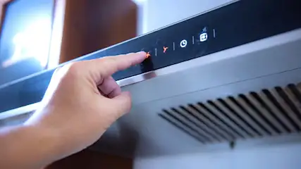 Een vinger drukt op een rood verlichte knop van een zwarte afzuigkap met ventilatierooster.