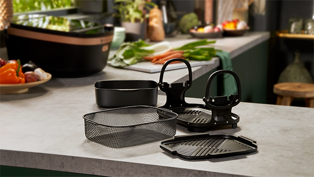 Keukenopstelling met airfryer accessoires, zoals een mandje, een bak en een rooster, en groenten op de achtergrond.