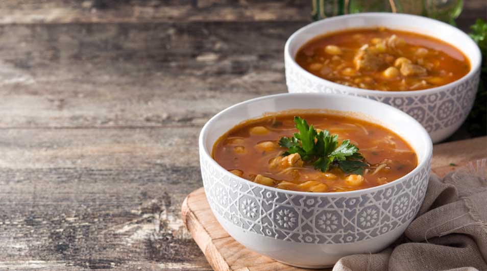 Twee kommen soep op een houten tafel. De kommen zijn wit met een grijs patroon en gevuld met oranje soep en garnering.