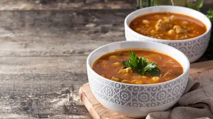 Twee kommen soep op een houten tafel. De kommen zijn wit met een grijs patroon en gevuld met oranje soep en garnering.