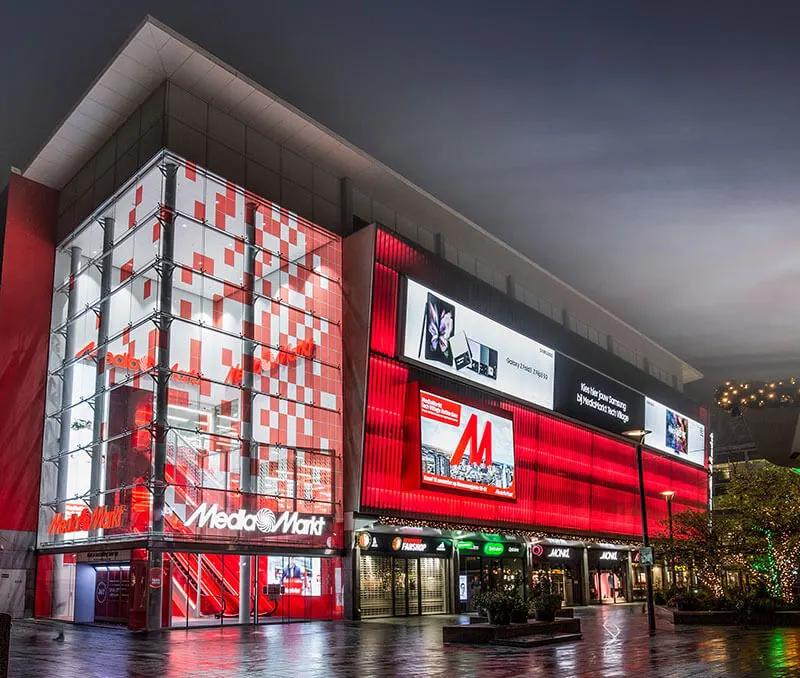 Groot, rood Media Markt gebouw met reclameborden en winkelpuien op een natte straat 's avonds.