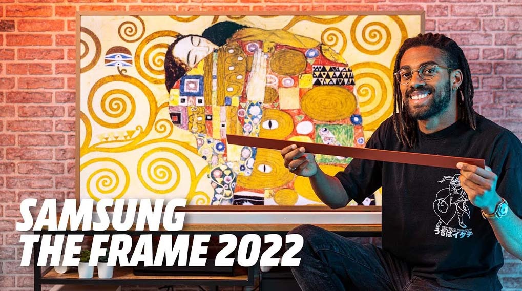 A person holds a frame piece in front of a framed painting with gold spirals on a brick wall. Text reads "Samsung The Frame 2022".