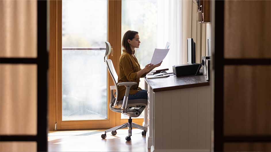 Vrouw zit aan bureau, leest papier. Bureau met printer en computer, witte stoel. Achtergrond met ramen en gordijnen.