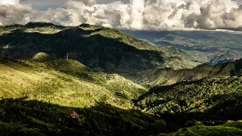 Heuvelachtig landschap met groene heuvels en bergen onder een bewolkte hemel. Een rode en witte toren staat op een heuveltop.