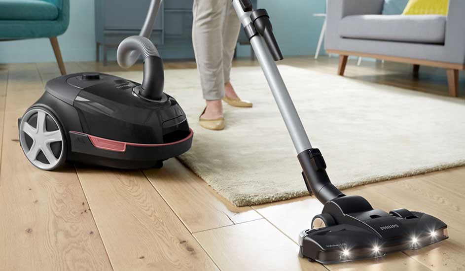 A person vacuums a wooden floor using a Philips vacuum cleaner, close to a rug and a sofa.
