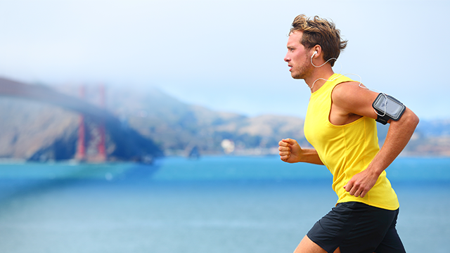 Een man in een geel shirt en zwarte korte broek rent met een telefoonhouder aan zijn arm. Achtergrond: blauw water en bergen.