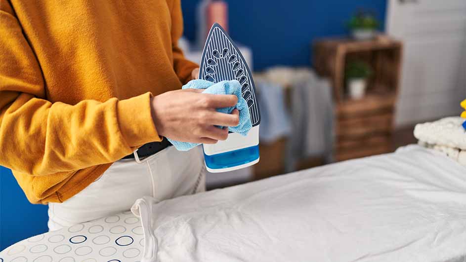 Iemand maakt een blauw-wit strijkijzer schoon met een blauwe doek op een strijkplank. Op de achtergrond staan opgevouwen handdoeken.