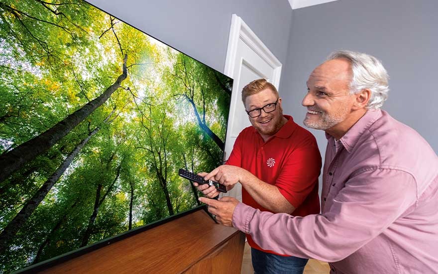 Een verkoper helpt een oudere man met het instellen van een grote, moderne televisie. Op het scherm staat een helder bos met groene bomen.